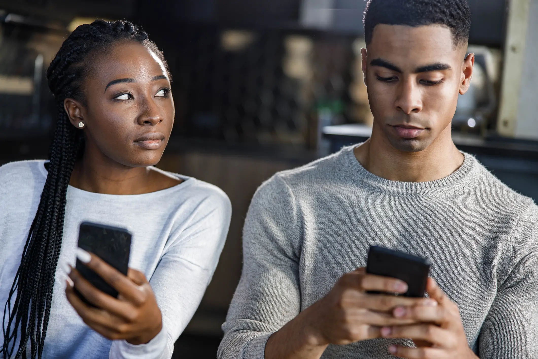A woman and man are seated next to each other on their phones. She is side eyeing him.